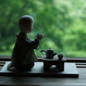 Una figurita de un monje arrodillado junto a una bandeja con una tetera y una taza, sobre un fondo borroso, verde y natural visto a través de una ventana.