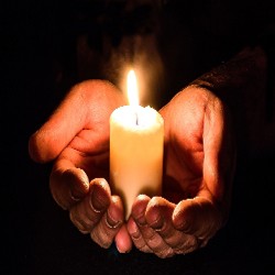 Dos manos ahuecan suavemente una vela encendida en la oscuridad, iluminando los dedos y proyectando un cálido y suave resplandor.